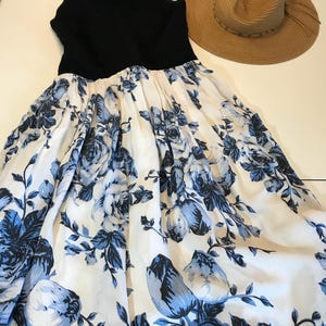 May include: A black sleeveless dress with a white skirt adorned with a blue floral design. A wide-brimmed straw hat is placed next to the dress. The dress has a beaded neckline.