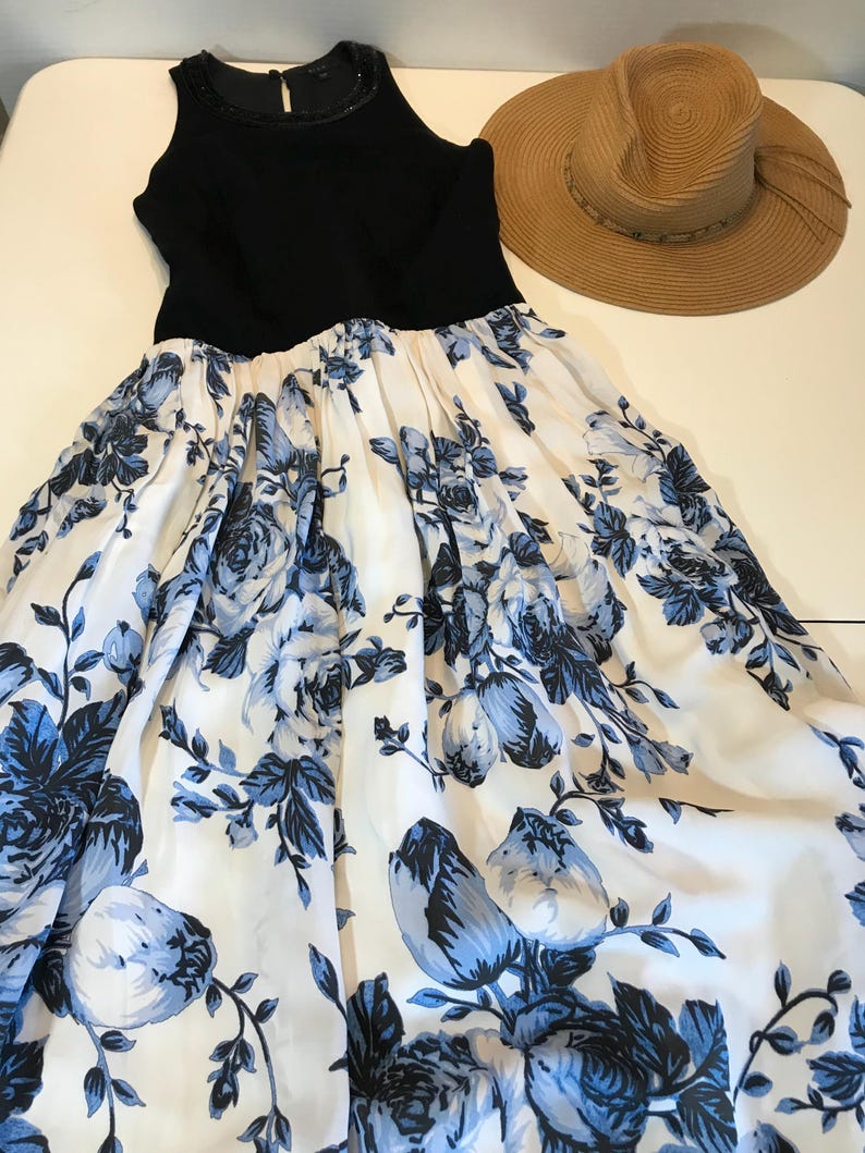 May include: A black sleeveless dress with a white skirt adorned with a blue floral design. A wide-brimmed straw hat is placed next to the dress. The dress has a beaded neckline.