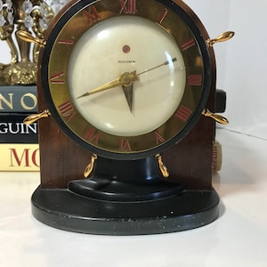 Könnte beinhalten: Eine Vintage-Telechron-Uhr mit goldenem Zifferblatt und römischen Ziffern. Die Uhr ist auf einem Holzsockel mit einem Schiffsrad-Design und goldenen Akzenten montiert.