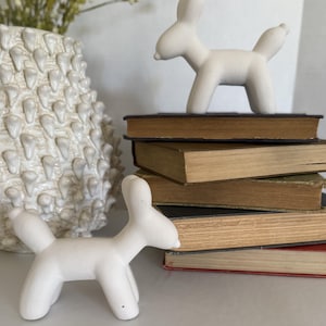 May include: Two matte white ceramic animal figurines shaped like balloon dogs, each featuring a smooth surface, four legs, and a rounded tail. One figurine stands on a stack of four vintage books, while the other stands on a flat surface nearby.