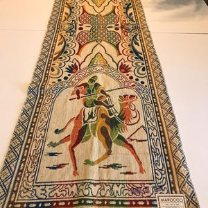 Op de afbeelding: Een rechthoekige, beige Marokkaanse tafelloper met een gedetailleerd, kleurrijk ontwerp. De centrale afbeelding toont een persoon die op een kameel rijdt. De loper heeft geometrische patronen, bloemmotieven en een geschulpte rand. Het label luidt "Marokko" en "Made in Italy".