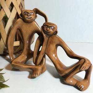 May include: Two brown ceramic monkey figurines with pink rhinestone eyes and accents. One monkey is kneeling, and the other is leaning. The figurines are displayed on a white surface, with a woven basket and greenery in the background.