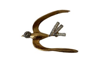 May include: A gold-toned bird brooch with a textured body and a head adorned with small, sparkling stones. The wings feature three rows of clear, glittering crystals, creating a striking contrast.