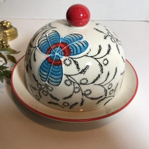 May include: A white ceramic butter dish with a red rim and a matching lid. The lid features a red knob and a floral design in blue, red, and black. The dish is designed for serving butter.