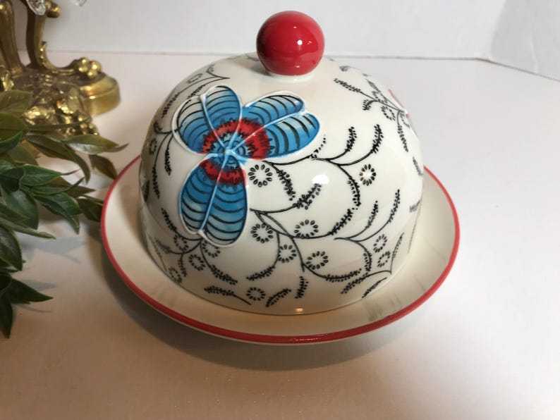 May include: A white ceramic butter dish with a red rim and a matching lid. The lid features a red knob and a floral design in blue, red, and black. The dish is designed for serving butter.