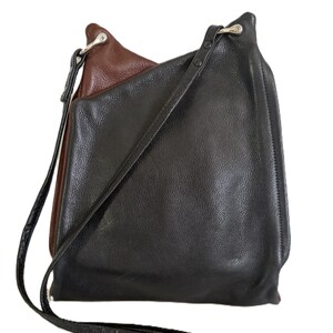 Puede incluir: Un bolso de hombro de cuero negro y marr&oacute;n con una correa larga. El bolso tiene un dise&ntilde;o en capas, con el cuero marr&oacute;n visible en la parte superior y el cuero negro formando el cuerpo principal. La correa es negra y parece ser ajustable.