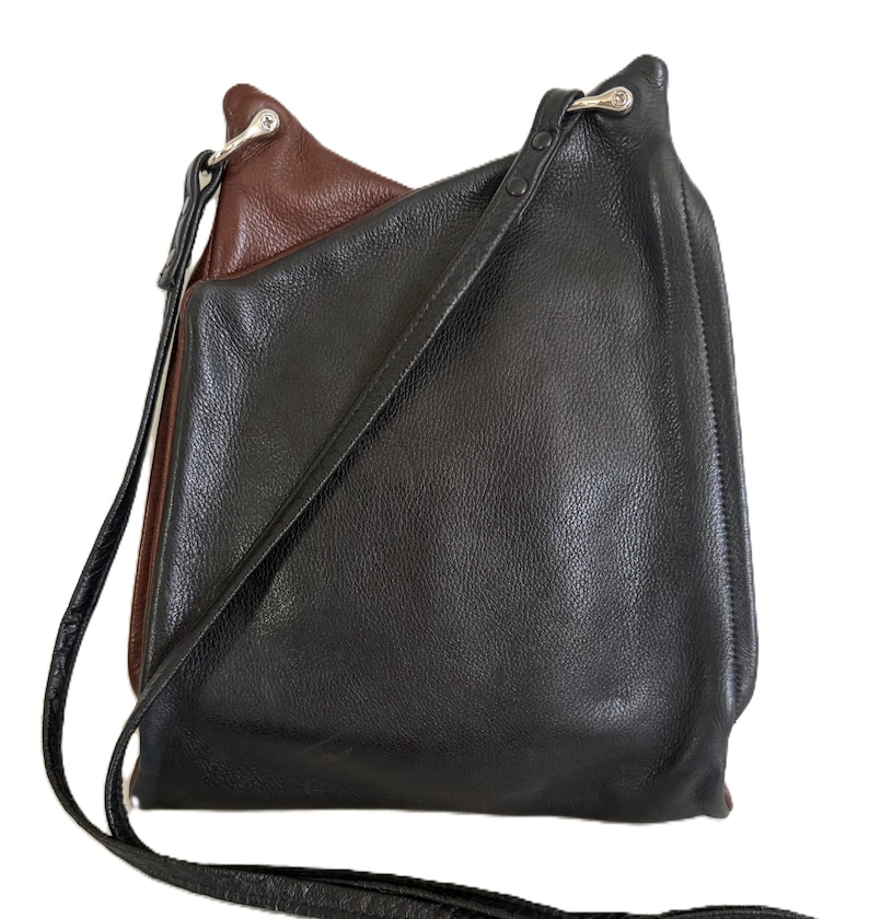 Puede incluir: Un bolso de hombro de cuero negro y marr&oacute;n con una correa larga. El bolso tiene un dise&ntilde;o en capas, con el cuero marr&oacute;n visible en la parte superior y el cuero negro formando el cuerpo principal. La correa es negra y parece ser ajustable.