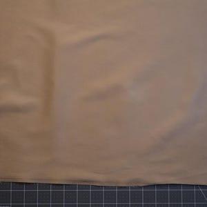 May include: A solid tan fabric piece laid flat against a dark gray cutting mat with white grid lines. The fabric has a smooth, matte texture and a straight, raw edge along the bottom, with the number 32 and 33 visible on the measuring grid.