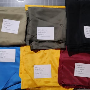 May include: Six folded fabric swatches in beige, olive green, black, bright blue, yellow, and deep red. Each piece of fabric features a white rectangular label with printed text detailing item codes, lengths in yards, and widths in inches.