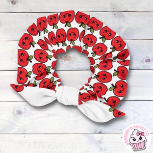 May include: A white headband with a repeating pattern of smiling red apples with green leaves. The headband is tied in a bow at the front. The background is a white wooden surface.