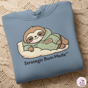 Könnte beinhalten: Hellblauer Hoodie mit einem Cartoon-Faultier, das einen grünen Hoodie trägt und auf einer Wolke liegt. Der Text "Strategic Bum Mode" ist darunter gedruckt.