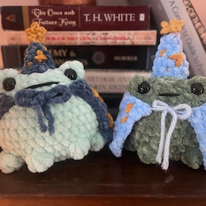 Puede incluir: Dos figuras de rana de peluche, una azul claro y otra verde, cada una con un sombrero y una capa de mago. Los sombreros son azules con estrellas doradas, y las capas son azules con detalles naranjas. Las ranas tienen ojos negros y una pajarita.