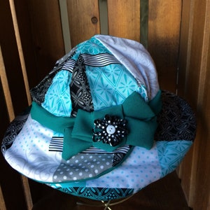May include: A turquoise and white patterned sun hat with a large black and white flower accent. The hat has a wide brim and is made of fabric.