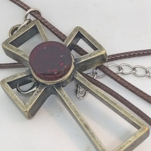 Könnte beinhalten: Ein goldfarbener Kreuzanhänger mit einem roten Glasstück in der Mitte. Der Anhänger ist an einer braunen Schnur und einer silbernen Kette befestigt.