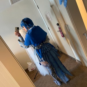 Peut inclure: Une personne portant un costume bleu et blanc avec une longue queue bleue. Le costume comprend une perruque blanche et une cape bleue. La personne tient un smartphone dans sa main droite.