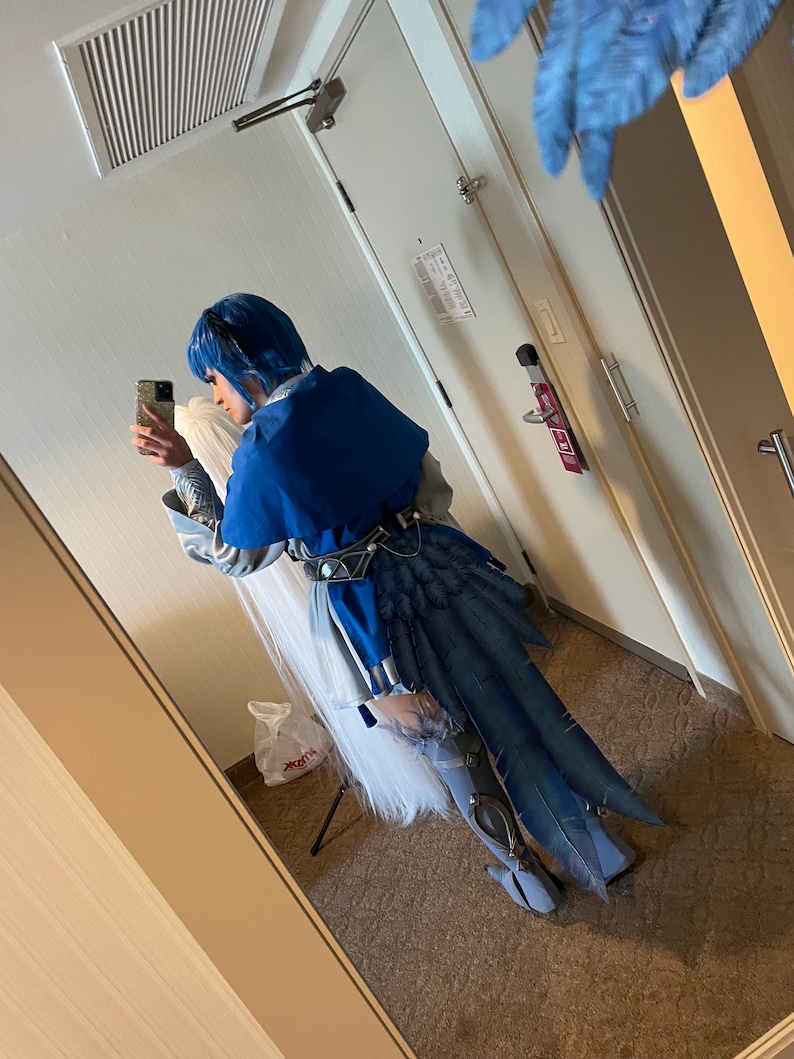 Peut inclure: Une personne portant un costume bleu et blanc avec une longue queue bleue. Le costume comprend une perruque blanche et une cape bleue. La personne tient un smartphone dans sa main droite.
