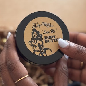 Könnte beinhalten: Ein schwarzer Körperbutterbehälter mit einem braunen Etikett, das eine Illustration einer Frau im Vintage-Stil und die Worte "Love Me" und "Body Butter" zeigt. Der Behälter wird von einer Person mit einem goldenen Ring und weißem Nagellack gehalten.