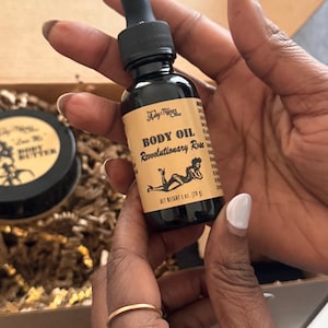 Può includere: Una bottiglia di vetro nero di olio per il corpo con contagocce, etichettata "Body Oil Revolutionary Rose". L'etichetta presenta una grafica di una donna. Il peso netto è di 28 g. Un barattolo di burro per il corpo è visibile sullo sfondo.