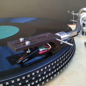 Puede incluir: Primer plano de un tocadiscos con un plato negro y un brazo de lectura con una cápsula de madera. El brazo de lectura está unido a una base de brazo de lectura plateada. El brazo de lectura está posicionado sobre un disco de vinilo negro con una etiqueta central verde. El brazo de lectura está equipado con una cápsula Ortofon.