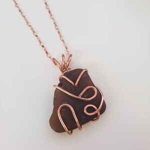 以下が含まれることがあります： 濃い茶色のシーグラスペンダントが付いた手作りのネックレス。ペンダントは銅線で包まれ、繊細な銅のチェーンから吊り下げられています。ネックレスは白い背景に表示されています。