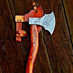 May include: A handmade axe with a Damascus steel head and a wooden handle. The handle is decorated with a carved design. The axe comes with a leather sheath.