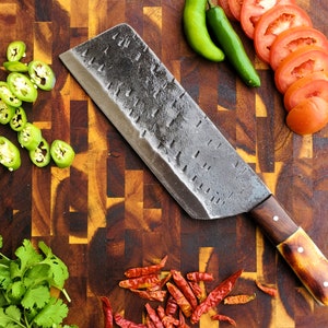 May include: A large, black, metal cleaver with a wooden handle sits on a wooden cutting board. Red and green peppers, and red tomatoes are also on the board.