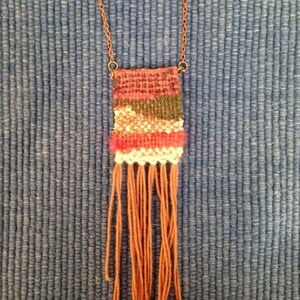 May include: A woven tapestry necklace with a brown chain and a rectangular tapestry with red, pink, white, green, and brown stripes. The tapestry has a fringe of brown yarn.