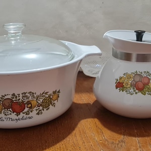 May include: White ceramic cookware set with a floral design featuring a casserole dish with a glass lid and a teapot with a black handle. The casserole dish has the text "La Marjolaine" on the bottom.