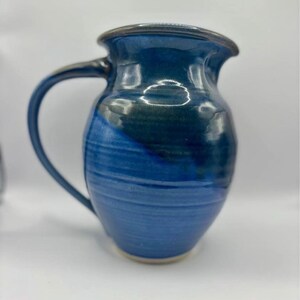 May include: A blue and green ceramic pitcher with a handle. The pitcher has a glazed finish and a rounded shape.