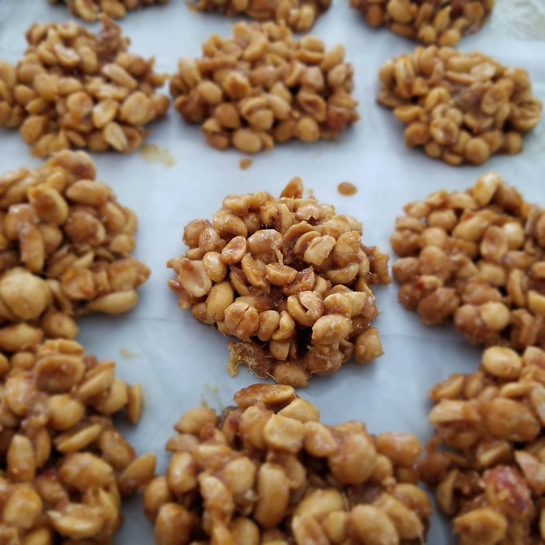 May include: A close-up of a baking sheet with peanut brittle. The brittle is made with peanuts and caramel and is arranged in small mounds on the baking sheet.