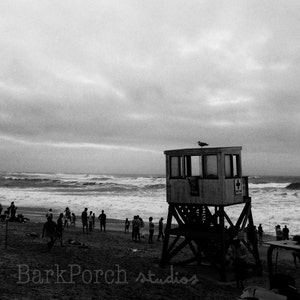 May include: A black and white photo of a lifeguard stand on a beach with people in the background. The stand has a red cross on it and a bird perched on the top.