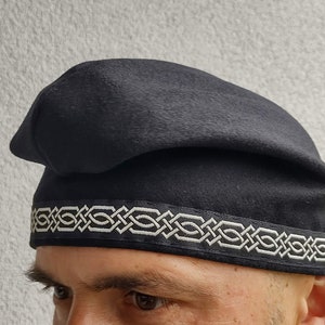 Op de afbeelding: Een zwarte wollen hoed met een witte gevlochten band. De band heeft een Keltisch knoopontwerp.