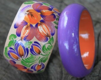 May include: Two wooden bangles, one painted with orange, purple, and green floral designs and the other solid purple with an orange interior.