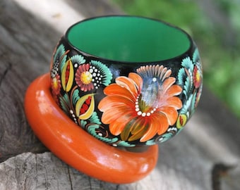 May include: A wooden bowl with a green interior and hand-painted floral designs in orange, red, yellow, and green. The bowl is sitting on a wooden base.