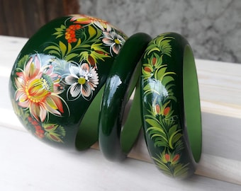 May include: Three green wooden bangles with floral designs painted in yellow, orange, white, and green. The bangles are stacked on top of each other.