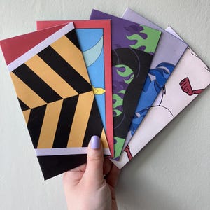 May include: A set of five rectangular paper envelopes fanned out, featuring distinct graphic designs including yellow and black diagonal stripes, a blue crescent moon, purple and green flames, blue hair patterns, and a light pink character illustration.