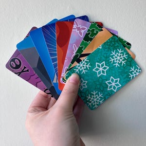 May include: A fan of rectangular playing cards featuring diverse designs including a purple card with a skull icon, a blue card with radial lines, a red card, a light purple card with white floral patterns, and a teal card with white snowflake motifs.