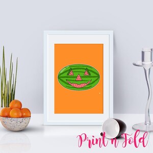 May include: A framed art print featuring a green watermelon with a carved jack-o'-lantern face on a solid orange background. The watermelon has triangular eyes and a nose, and a wide, jagged mouth, all revealing the pink fruit flesh inside.