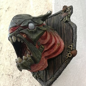 May include: A green and red monster head sculpture mounted on a wooden plaque with a textured, coral-like border. The monster has a large, open mouth with sharp teeth and a single, white eye.