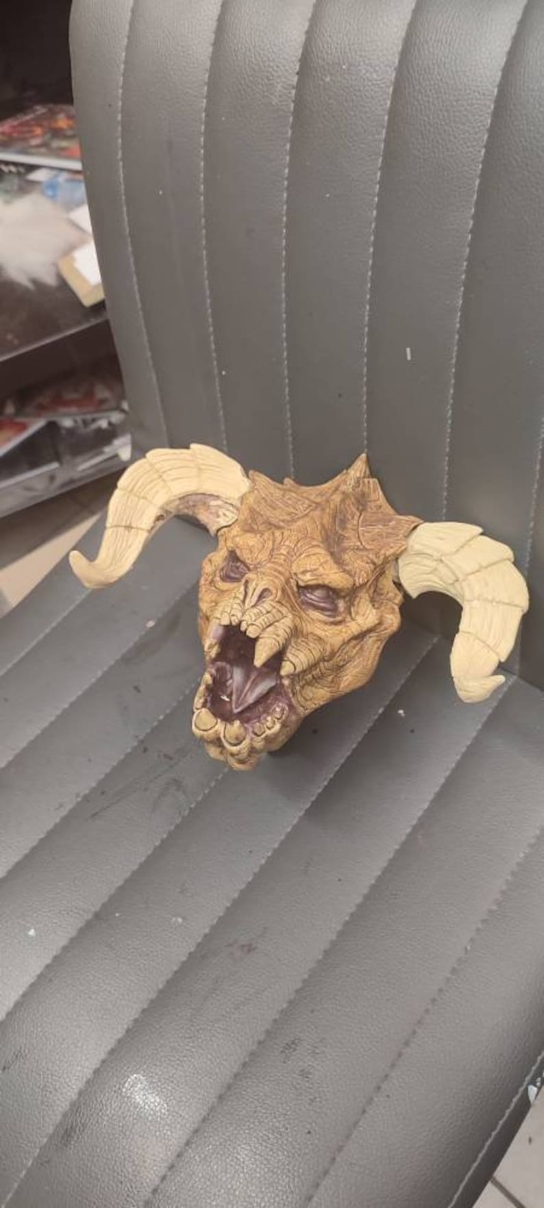 May include: A brown and tan monster mask with horns. The mask has a wide open mouth with sharp teeth and a textured surface.