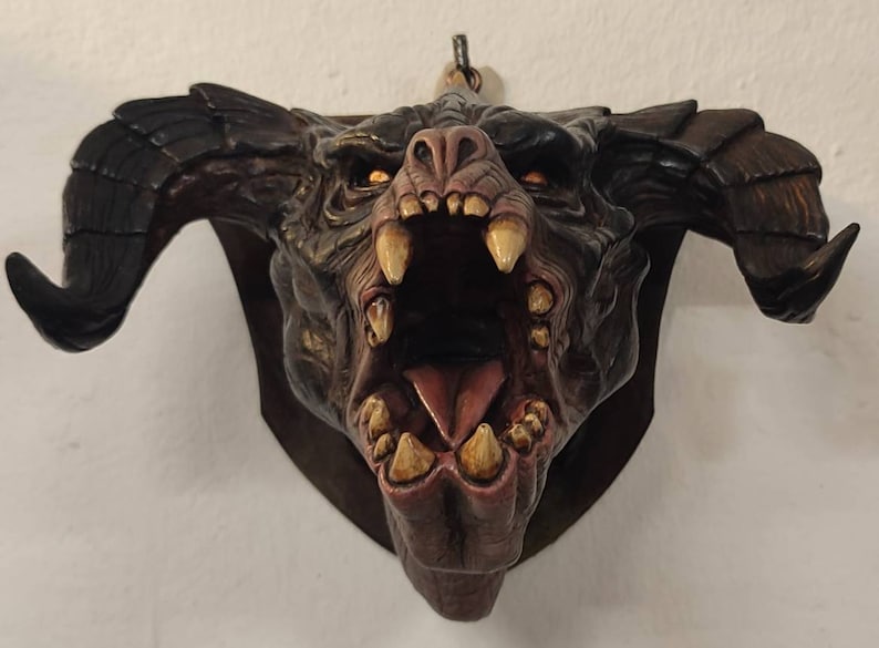 May include: A snarling, red and black monster head sculpture with large horns. The sculpture is mounted on a wooden plaque and has a rope hanging from the top.