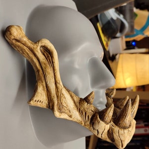 May include: A detailed, tan-colored jaw piece with sharp, conical teeth and a textured surface. The jaw is positioned on a gray mannequin head, showcasing its design and form. The piece appears to be a prop or costume element.