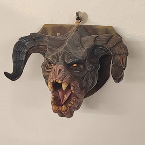 May include: A brown and black demon head sculpture with large horns and a wide open mouth, mounted on a wooden plaque.