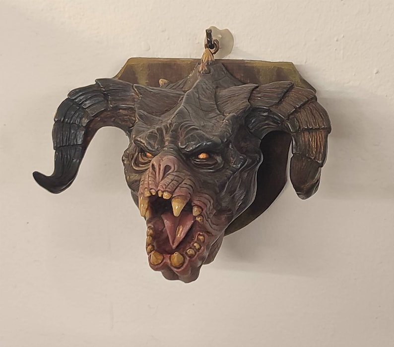 May include: A brown and black demon head sculpture with large horns and a wide open mouth, mounted on a wooden plaque.