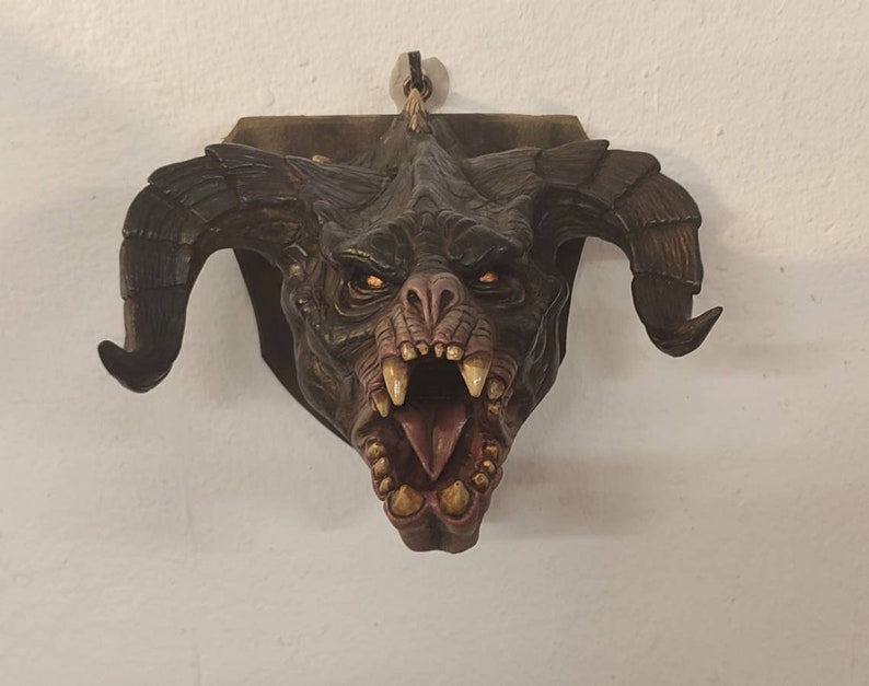 May include: A detailed, sculpted, wall-mounted, demonic creature head with large horns, a wide open mouth, and sharp teeth. The creature is painted in shades of brown, black, and red.