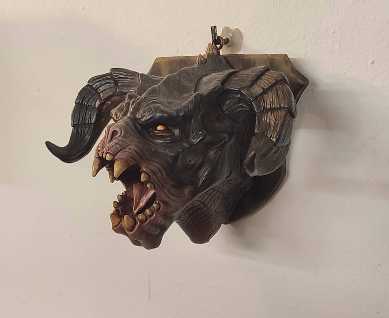 May include: A dark brown and black sculpted monster head with a toothy grin and large horns. The head is mounted on a wooden plaque.