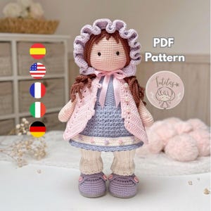 Puede incluir: Una muñeca de ganchillo hecha a mano, con cabello castaño, que lleva un gorro lavanda, una chaqueta rosa y un vestido azul. La muñeca tiene polainas color crema y zapatos lavanda. La imagen incluye el texto "PDF Pattern" y un logotipo.