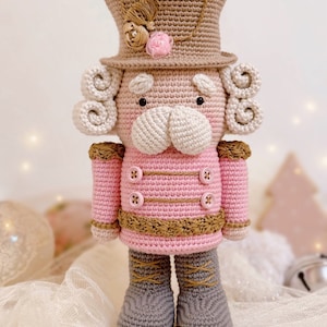 May include: A crocheted nutcracker doll with a pink jacket, brown hat, and gray boots. The doll has a white face with a brown mustache and a pink flower on its hat.