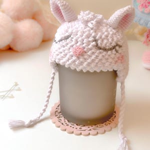 K&ouml;nnte beinhalten: Wei&szlig;er H&auml;kel-Babyhut mit Hasenohren und rosa gestickten Details. Der Hut sitzt auf einem rosa Spitzenuntersetzer.