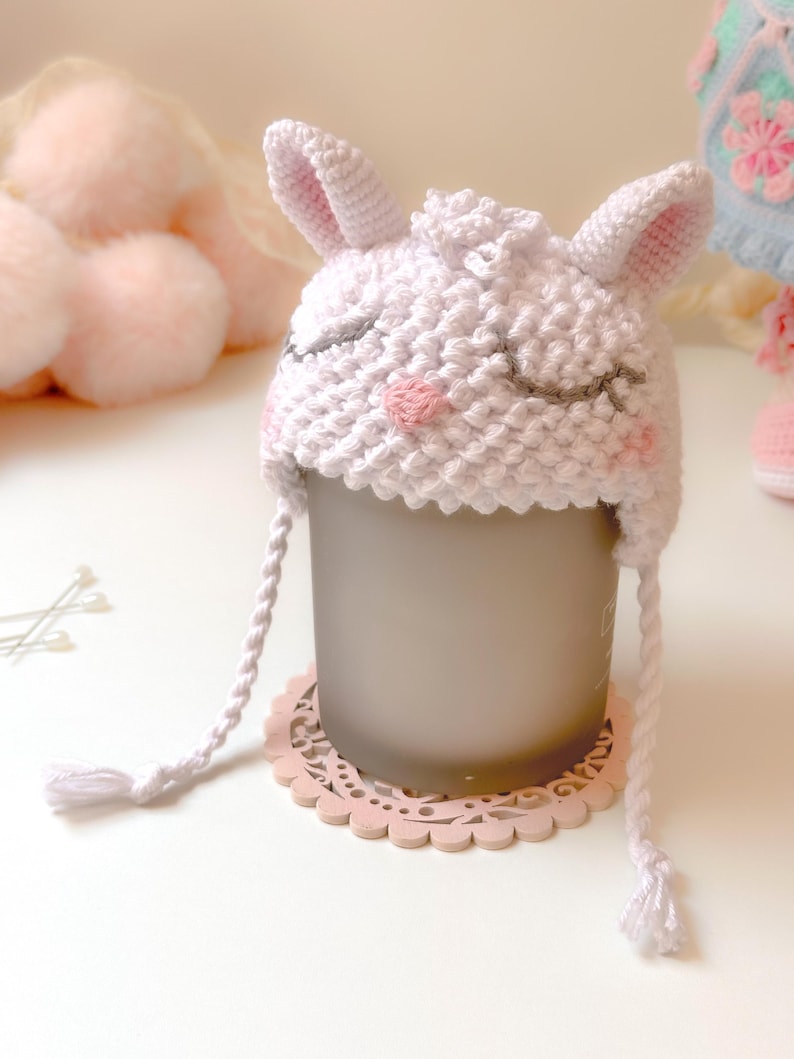 K&ouml;nnte beinhalten: Wei&szlig;er H&auml;kel-Babyhut mit Hasenohren und rosa gestickten Details. Der Hut sitzt auf einem rosa Spitzenuntersetzer.